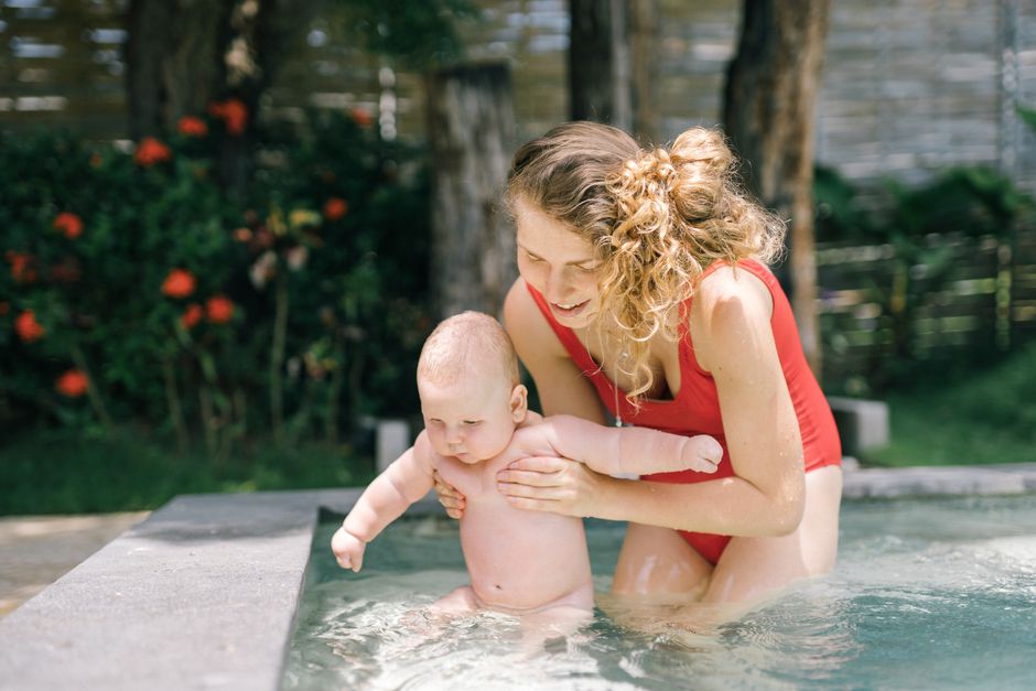 Mother supporting her baby in an outdoor swimming pool during summer, surrounded by greenery