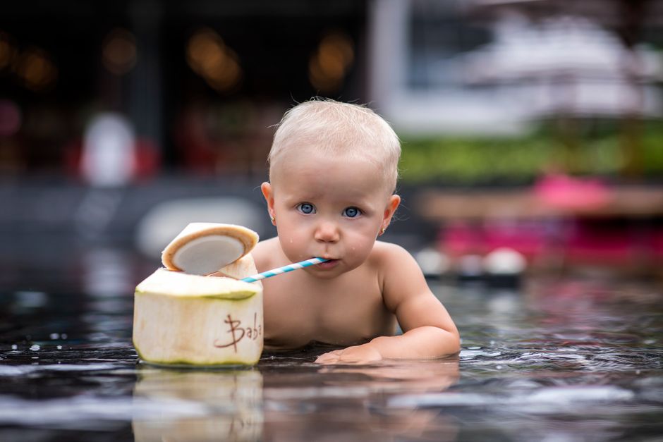 A baby with blue eyes enjoying coconut water through a straw in an outdoor pool