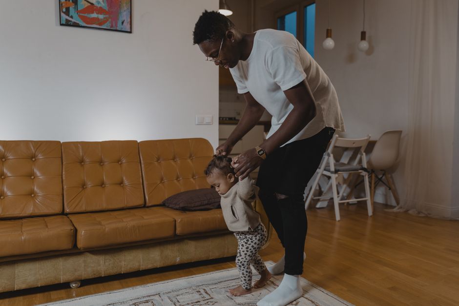 A father lovingly guides his toddler in taking first steps in the comfort of home