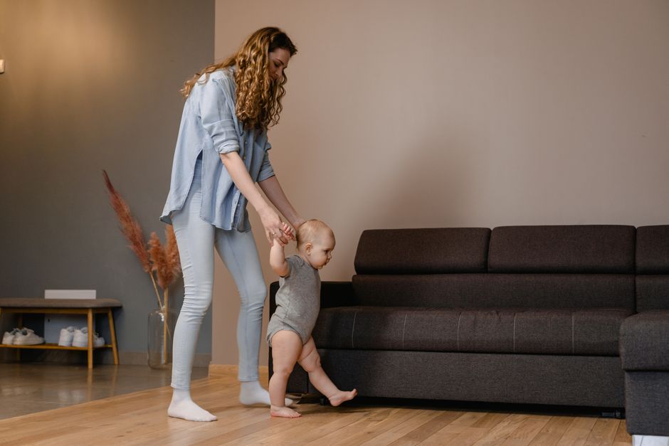 A mother supports her baby as they take first steps near a sofa indoors
