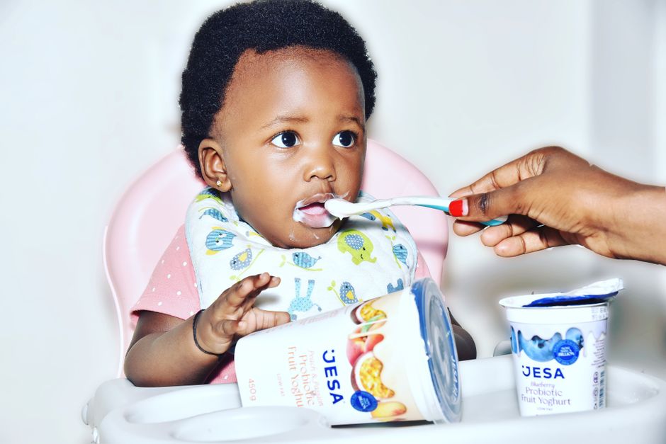 Baby in high chair being fed yogurt by a parent. Smiling with delight during snack time.