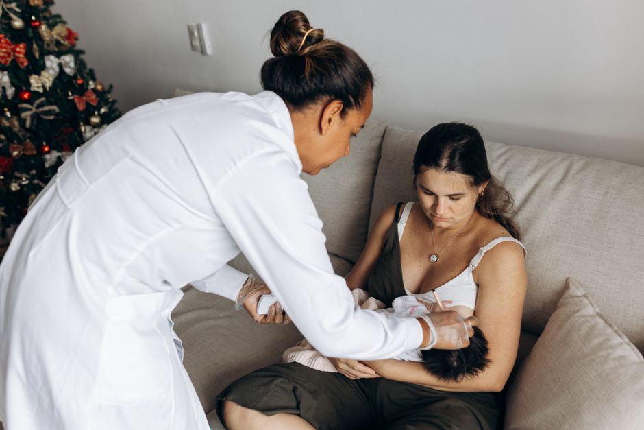 A healthcare professional helps a new mother care for her baby on a sofa during a home visit