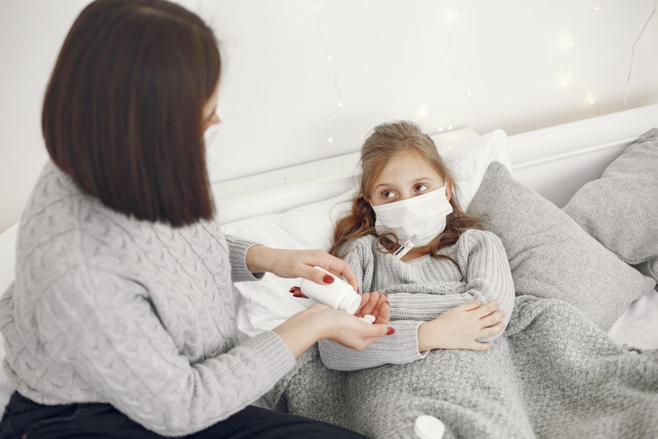A mother helps her sick daughter wearing a face mask in bed, offering medicine.