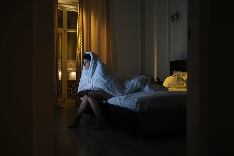 Woman wrapped in blanket watching screen, battling insomnia in dimly lit bedroom