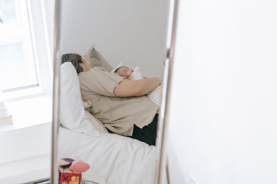 Anonymous female with sleeping infant baby in arms reflecting in mirror while resting on comfortable bed in light bedroom at home