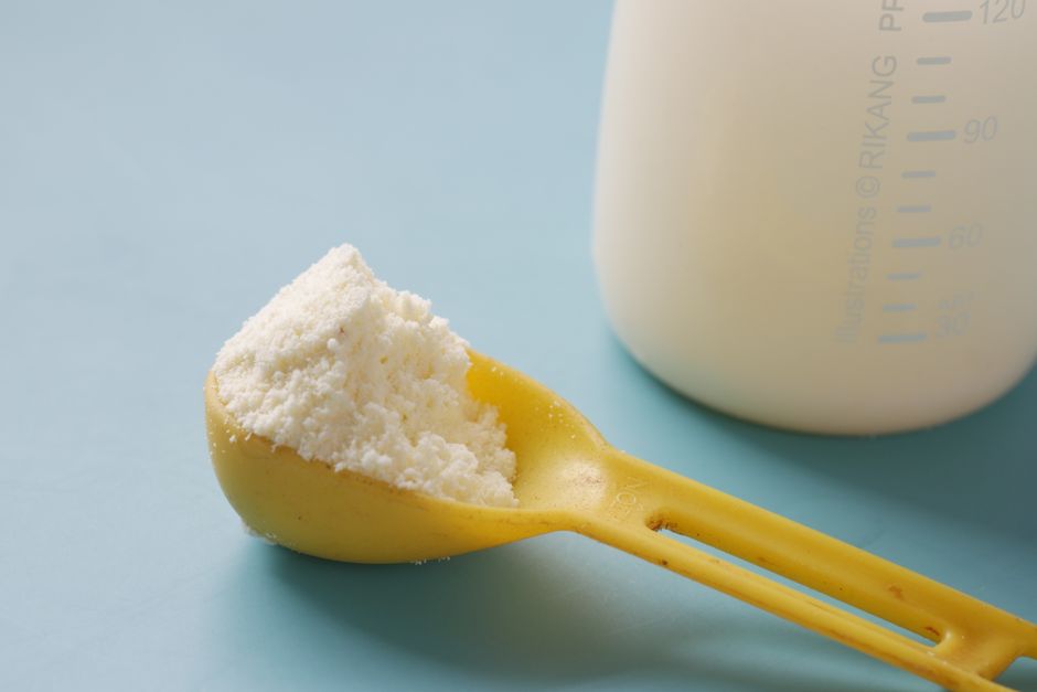 A yellow spoon filled with powdered milk, next to a measuring bottle, on a blue background