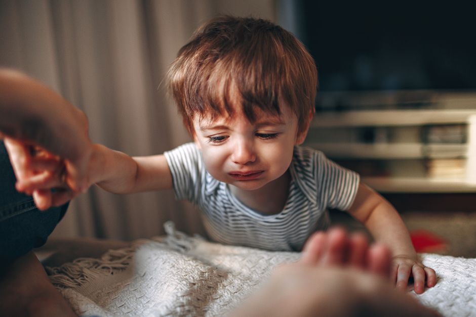 A close-up of an upset toddler reaching out while crying indoors