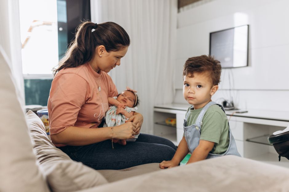 A mother sitting on a sofa with her baby and young son in a bright living room setting