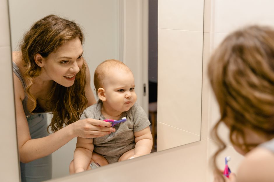 Mother helping baby brush teeth at bathroom mirror