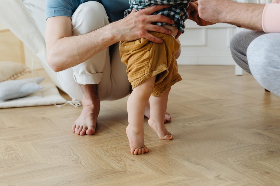 A heartwarming scene of a father helping his baby take first steps barefoot on a wooden floor