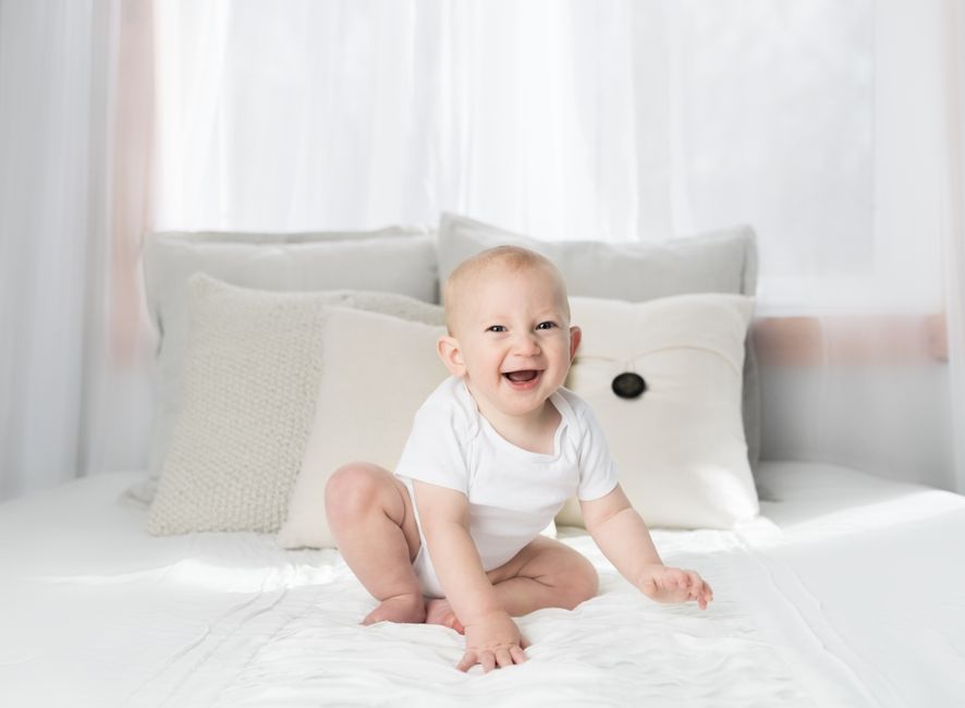 Cute baby sitting and smiling on a white bed in a bright, airy bedroom